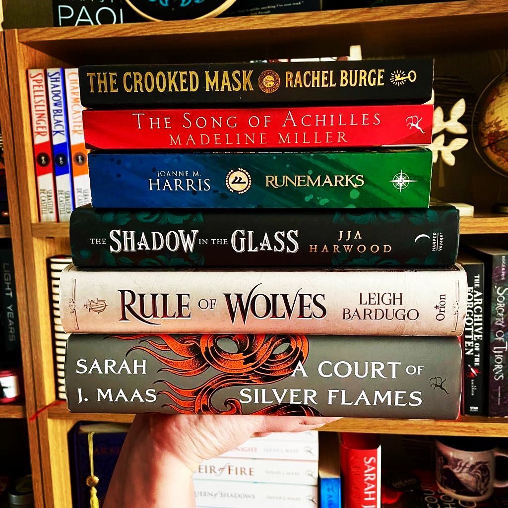 A stack of books including, The Crooked Mask by Rachel Burge, The Song of Achilles by Madeline Miller, Runemarks by Joanne M. Harris, The Shadow in the Glass by JJA Harwood, Rule of Wolves by Leigh Bardugo and A Court of Silver Flames by Sarah J. Maas 